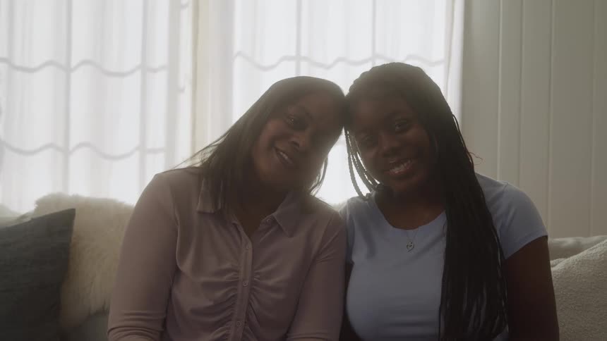 Two black females are sitting on a couch leaning head to head with each other smiling