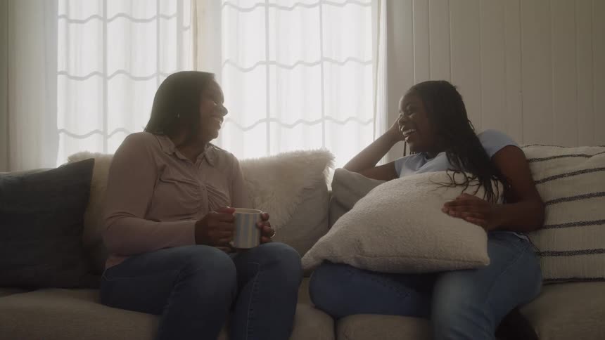 Two women are sitting on a sofa smiling as they chat and one of them is not holding a cushion