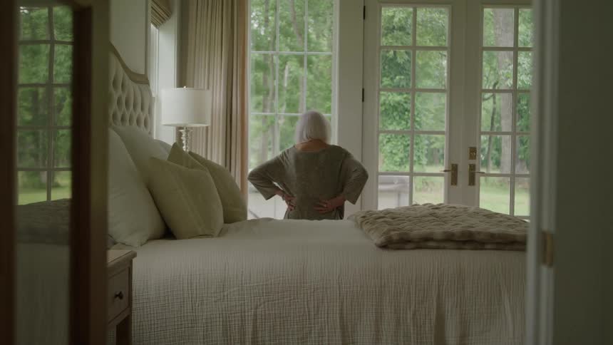 A woman looks out her tall bedroom windows