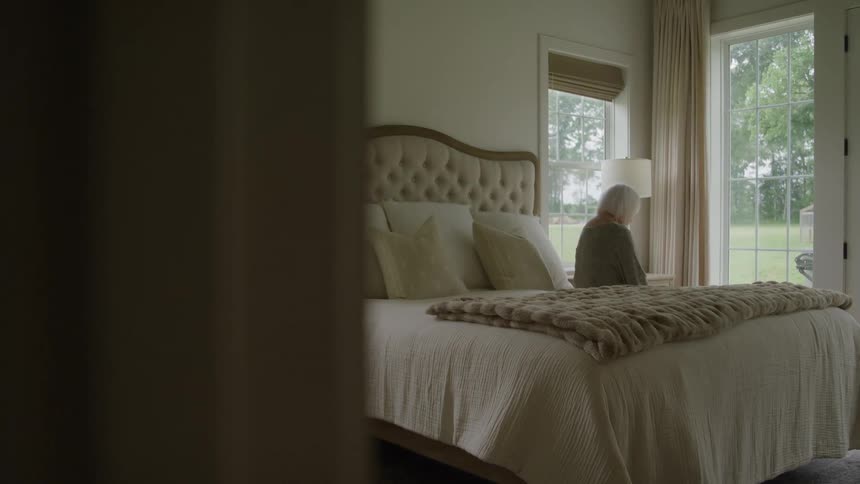 An elderly person sits on a bed looking out a window in a sunlit bedroom