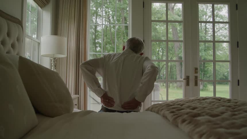 An elderly man sitting on the edge of a bed rubbing his aching lower back