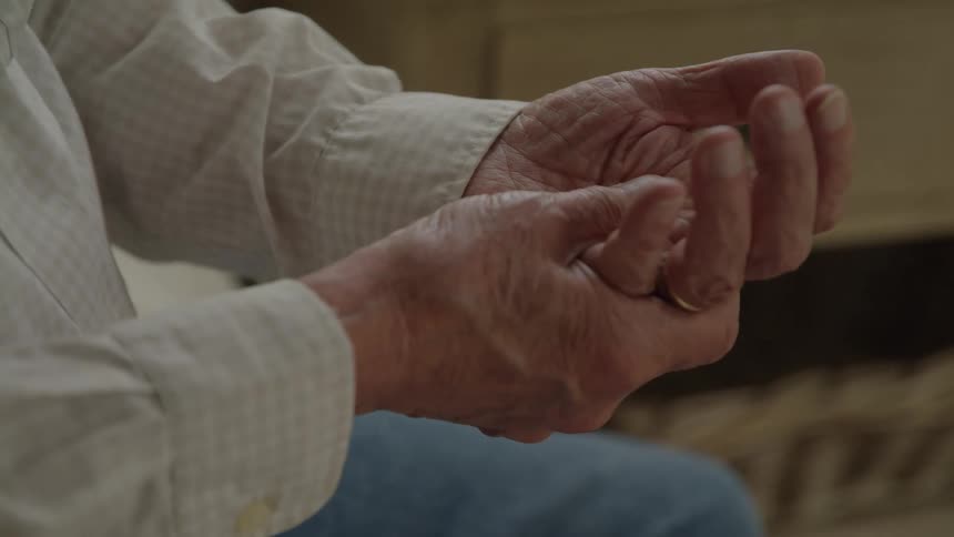 The video shows a closeup of an elderly persons hands which are clasped together in a gentle almost prayerlike position the hands appear wrinkled and aged and there is a simple ring visible on one of the fingers as they slowly open and close