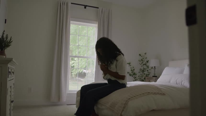 Woman sitting on the foot of her bed during the day prays