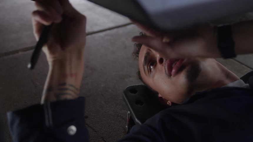 A focused male mechanic wearing dark work clothes and lying on a creeper is shown from below working on the underside of a vehicle with a tool in hand with his face turned slightly toward the light