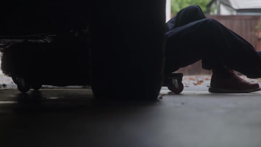 A mechanic lying on his back on a wheeled creeper is seen from a low angle as he slides out from underneath the shadowy rear of a vehicle with his brown work boots emerging into the daylight