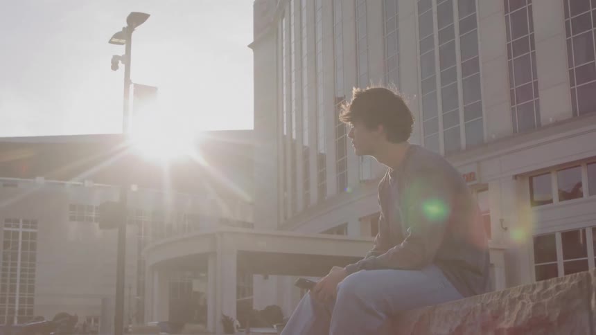 A teenage sits outside a modern building at sunset looking at his phone