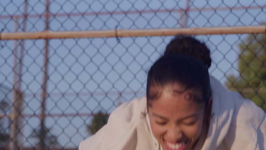 A young woman in a white hoodie smiles and laughs while crouching in an athletic stance in front of a chainlink fence