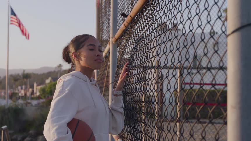 A young woman in a white coat holding a basketball has her hand on the fence of a basketball court looking at it