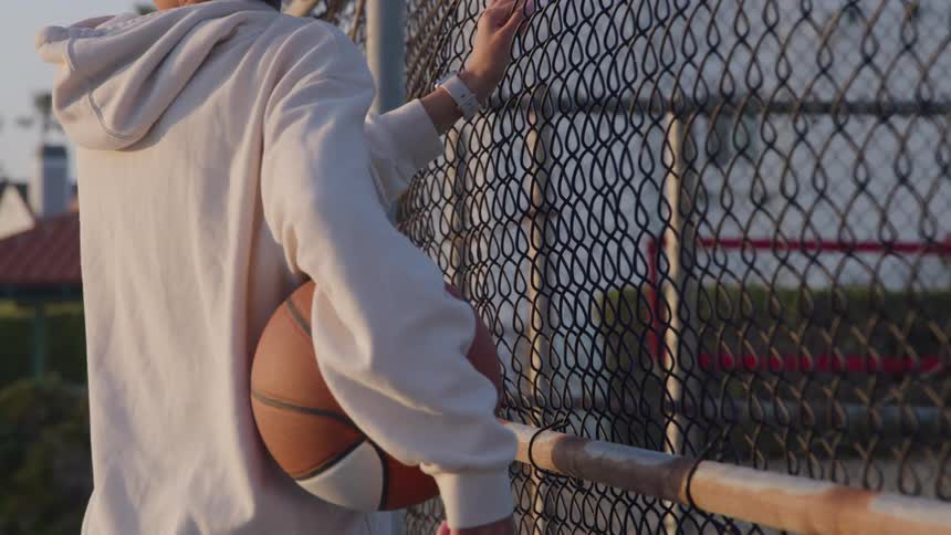A person in a white hoodie holds a basketball while resting against a chain link fence at an outdoor court