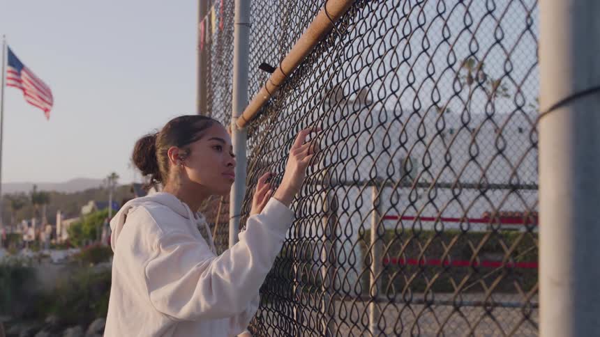 A woman in a white hoodie stands by a chainlink fence gazing thoughtfully through it with an american flag and palm trees visible in the background