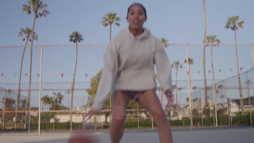 A young woman playing basketball by herself on an open court surrounded by palm trees