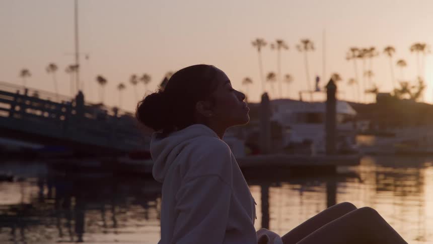 A young woman rests sitting downby a body of water near a pier with a docked boat