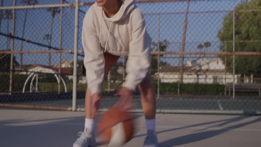 A young woman playing basketball by herself on an open court surrounded by palm trees