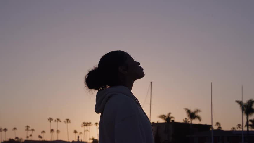 A woman in a hoodie looks upward as the sun sets behind her with palm trees and sailboat masts silhouetted in the background