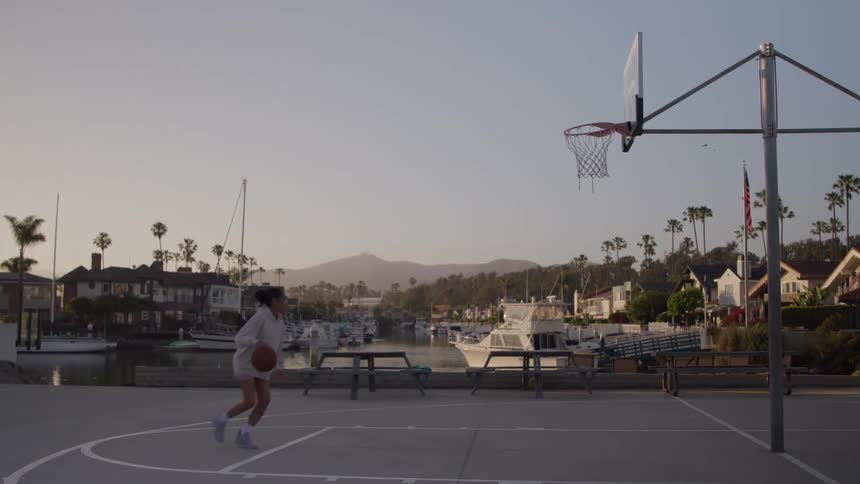A woman is playing basketball and dunking on a basketball court and behind her there is a marina full of boats