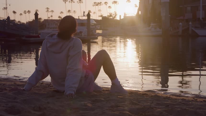 Woman sitting on the sand watching the sunset by the water