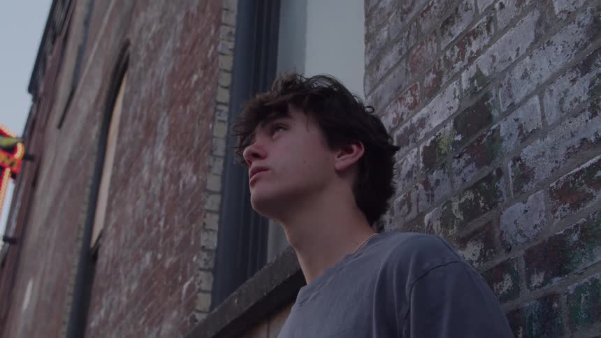 A young man with messy hair leans against a brick wall and looks up thoughtfully