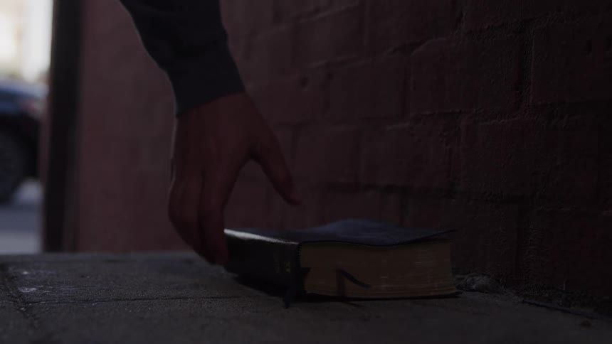 Hands of a white person picking up a black book from the floor on an alley next to the sidewalk of an urban area where other people are walking outdoors by day time