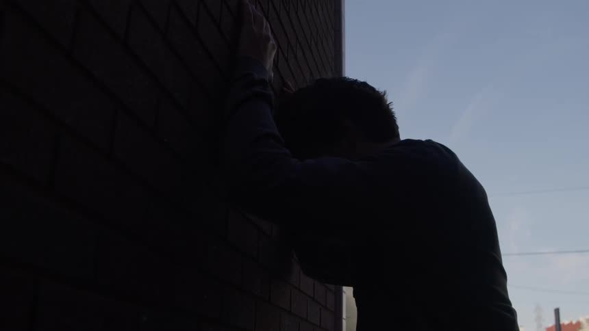 Silhouetted against the sky a distressed young man leans against a dark brick wall in an urban alley
