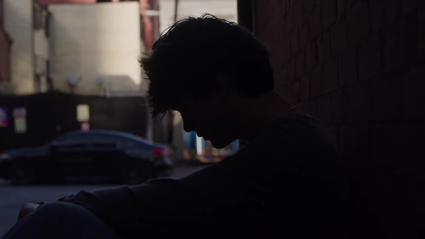 Teenage boy sits next to a building as cars and pedestrians pass by on the street