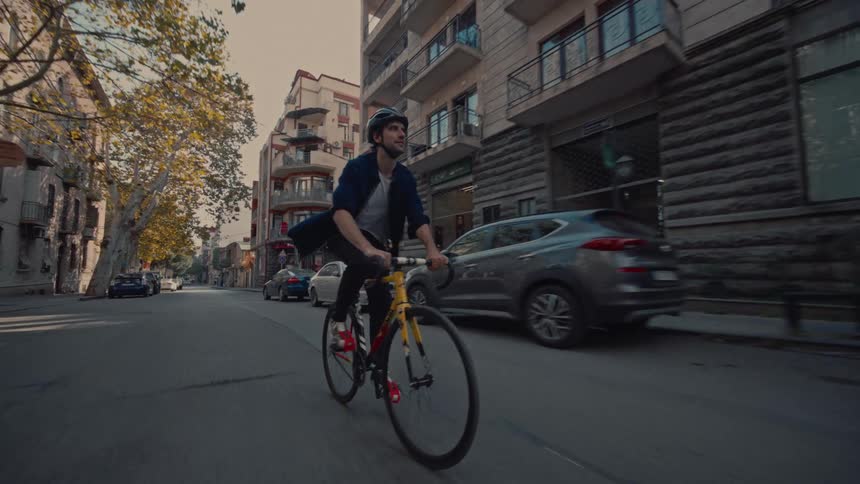 A young man wearing a helmet and a dark jacket is captured from a low angle as he pedals a distinctive yellow and black road bike down a wide urban street passing parked and moving vehicles with classic multistory buildings forming the backdrop