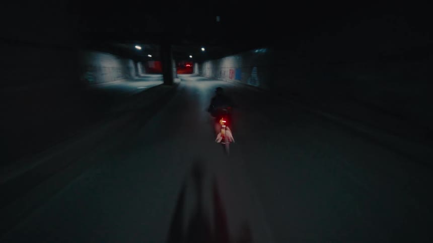 This video filmed from behind shows a person wearing a helmet and a dark top as they ride a bicycle through a dimly lit road the video begins as the cyclist is moving quickly through a tunnel or underpass with concrete walls covered in faded graffiti