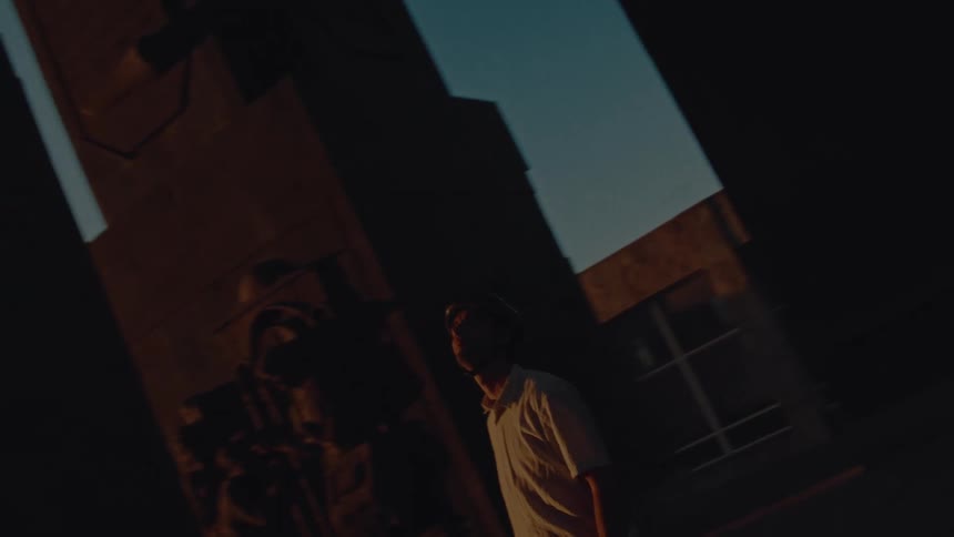 A person in a lightcolored polo shirt is dramatically framed in a shaft of sunlight against a mostly dark urban backdrop of tall buildings they look up and around as they slowly move into the shadows suggesting a journey or contemplation