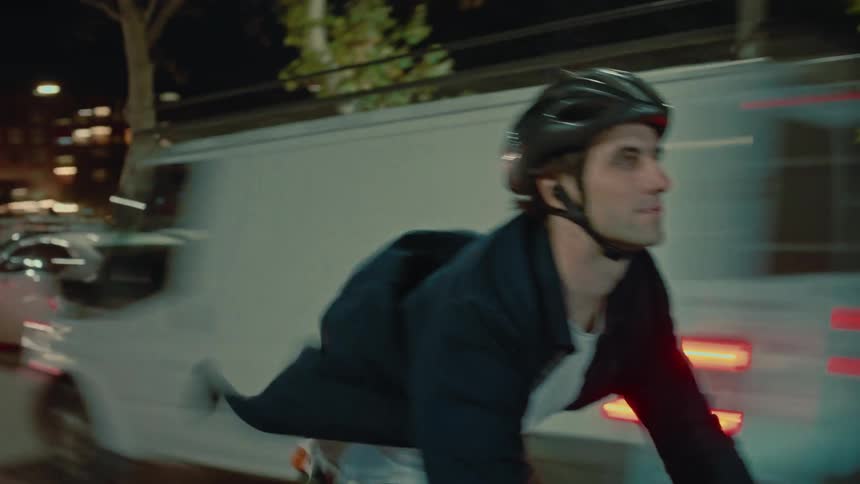 A cyclist wearing a helmet rides through a city at night passing traffic with many people walking in the background