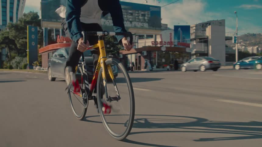 A focused male cyclist wearing a helmet and a dark jacket rides his distinct yellow and black road bike quickly along a sunny multilane city road maintaining speed as vehicles pass him and modern highrise buildings form the back of the scene