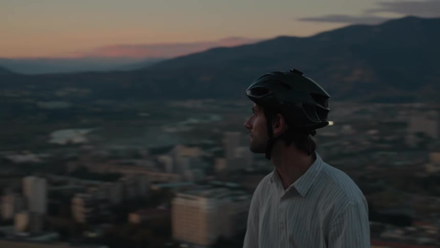 Man stands on a hill and looks at the city as red tints the sky above the nearby mountains