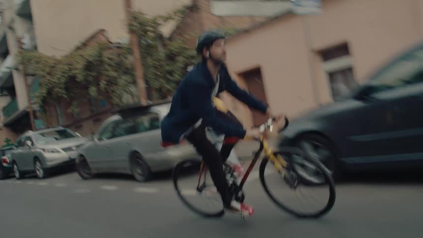 A cyclist wearing a helmet rides a yellow bike down a city street lined with parked cars and buildings passing by a few people on the sidewalk