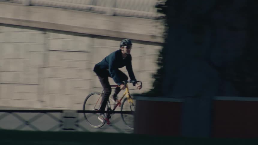 The video features a cyclist dressed in dark clothing and a helmet riding a distinctive yellow and red bicycle along the curb of a busy street with an overpass visible above he appears to be moving quickly as he passes signs and some shrubbery