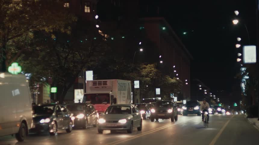 A long line of cars with bright headlights and taillights moves slowly along a wide urban street at night while a single cyclist rides past them on the right all illuminated by scattered streetlights and the windows of dark city buildings