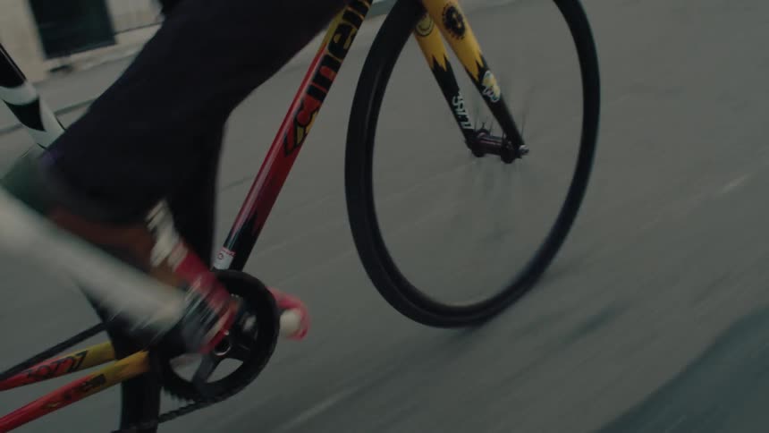 A cyclist wearing a helmet is rapidly pedaling a colorful trackstyle bicycle down an urban street and then transitions to a more open road