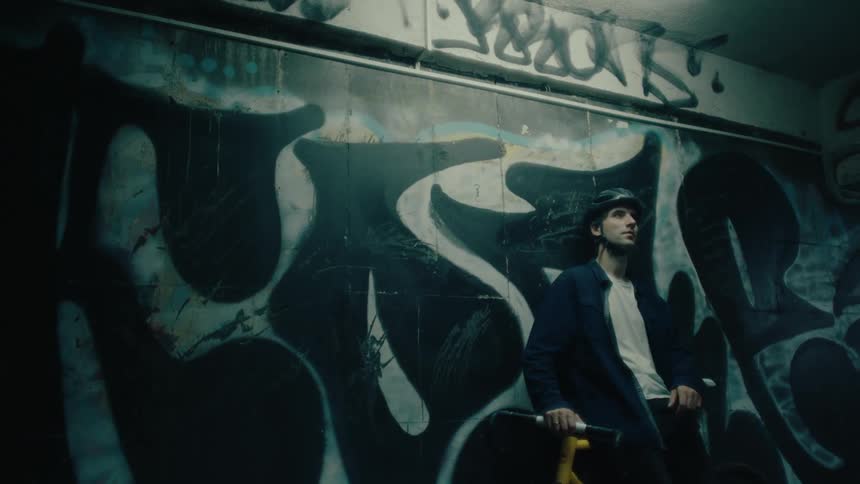 A cyclist wearing a helmet leans against his bike in a dimly lit tunnel covered with graffiti as people walk by
