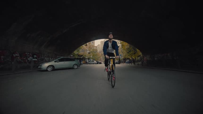Male cyclist pedals along a highway passing apartment buildings and under a bridge on an autumn day