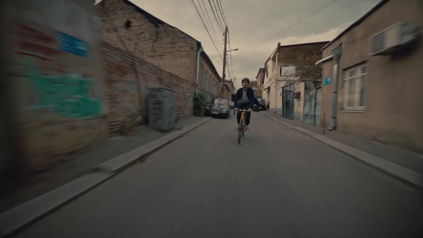 A man wearing a helmet is riding a bicycle on an asphalt street with some cars parked on either side