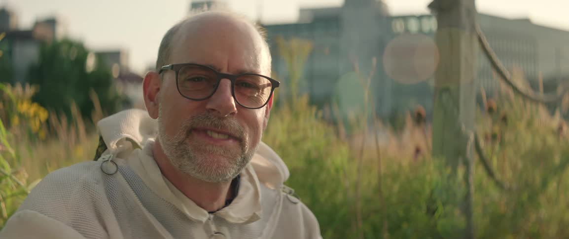 A middleaged beekeeper is smiling gently and looking directly into the camera near his hives