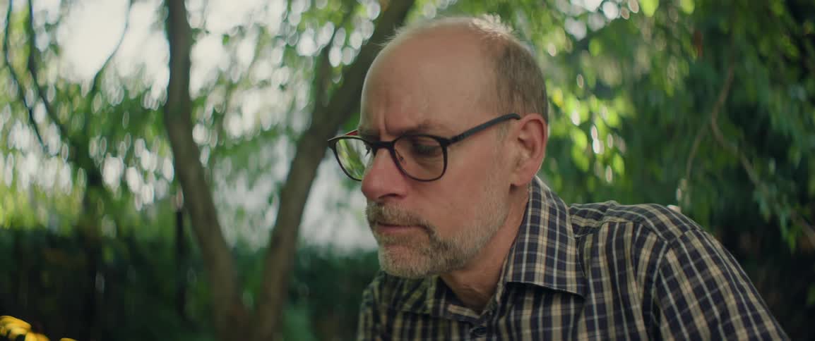 Man with glasses looks intently at something in an outdoor garden setting