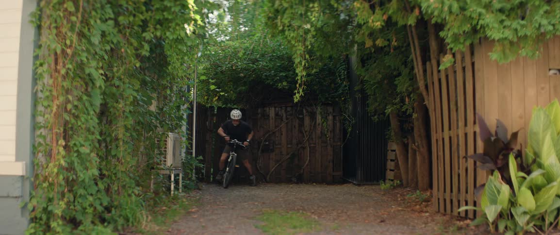 A man wearing a bicycle helmet on a bike emerges from an overgrown garden gate