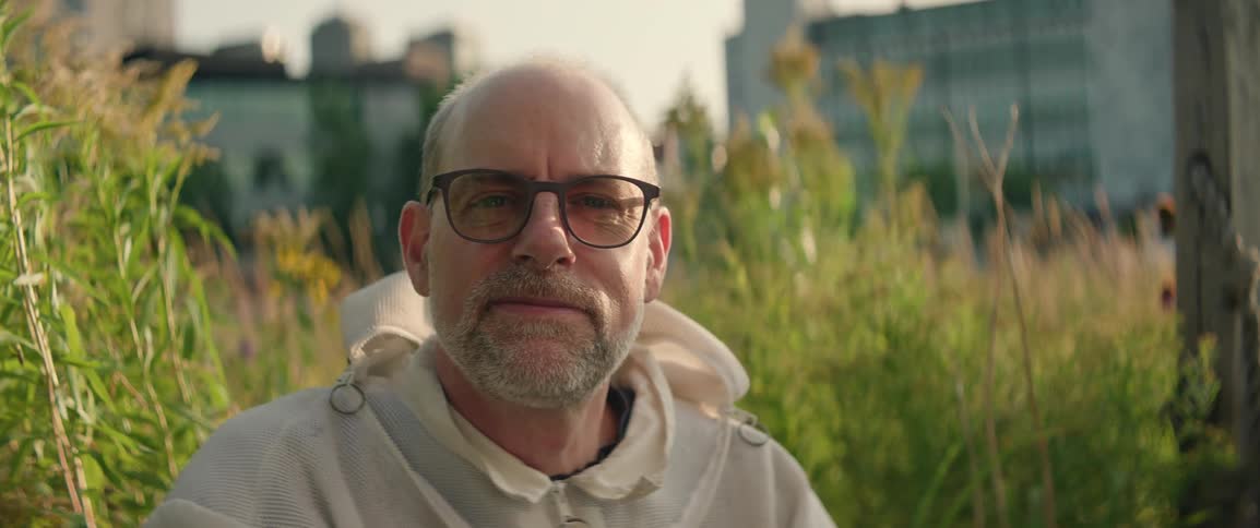 Beekeeper wearing glasses and protective clothing smiles amid vegetation