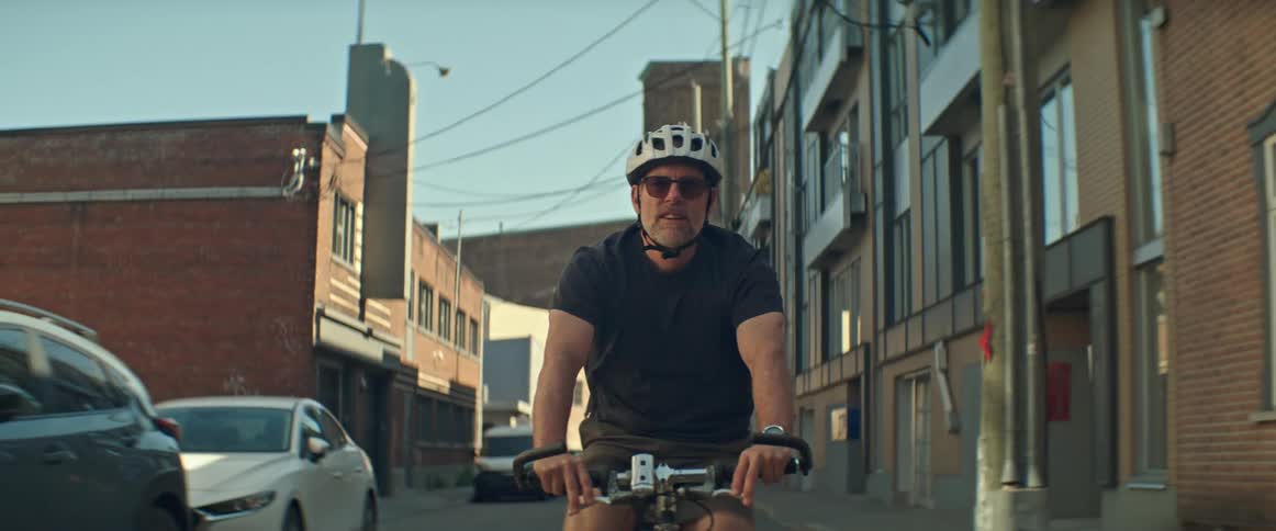 A man wearing a helmet and sunglasses rides his bicycle down a narrow urban street lined with brick and industrial buildings