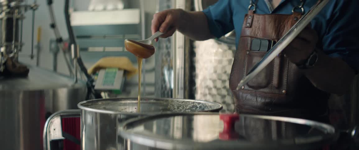 Man wearing a leather apron pours liquid honey into a metal container