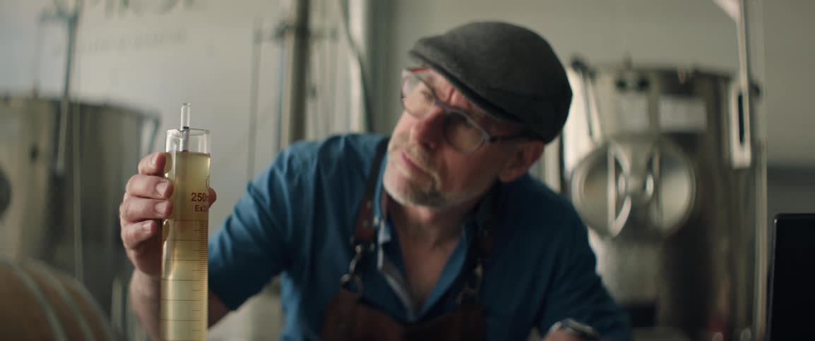 A man wearing a leather apron and blue shirt works carefully with a tall glass cylinder filled with liquid in a workshop