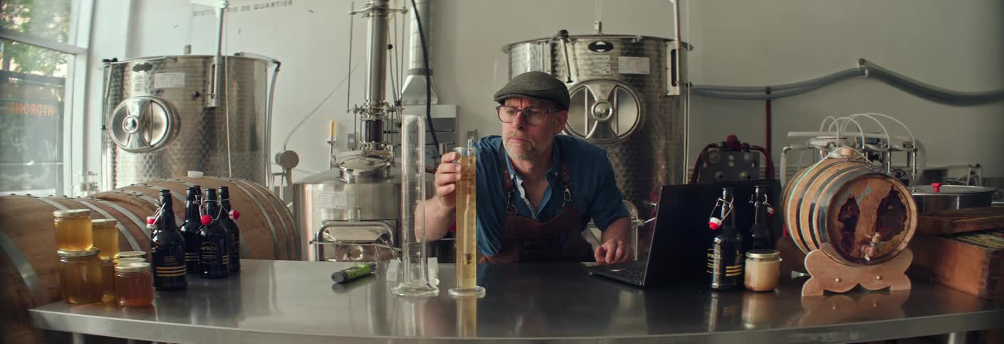 A distiller wearing a cap and a brown leather apron is meticulously conducting a liquid test using a hydrometer