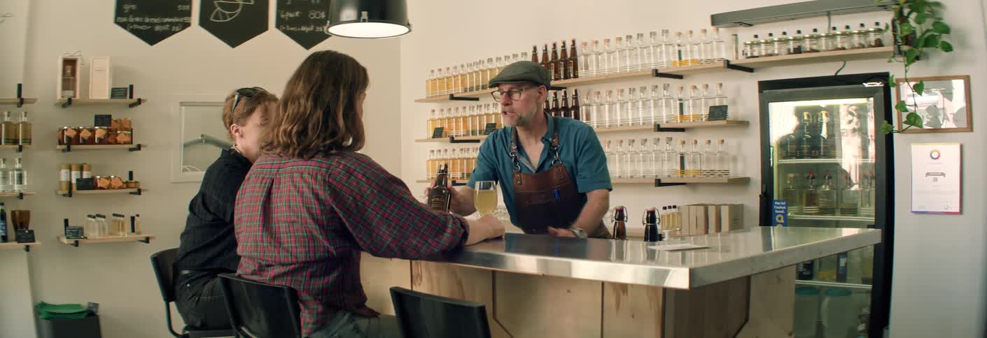 A bartender wearing an apron and a cap is standing behind a metal counter presenting a dark bottle to two seated female customers