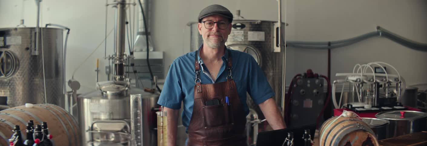 Man wearing a leather apron and cap poses in a distillery surrounded by equipment and bottles