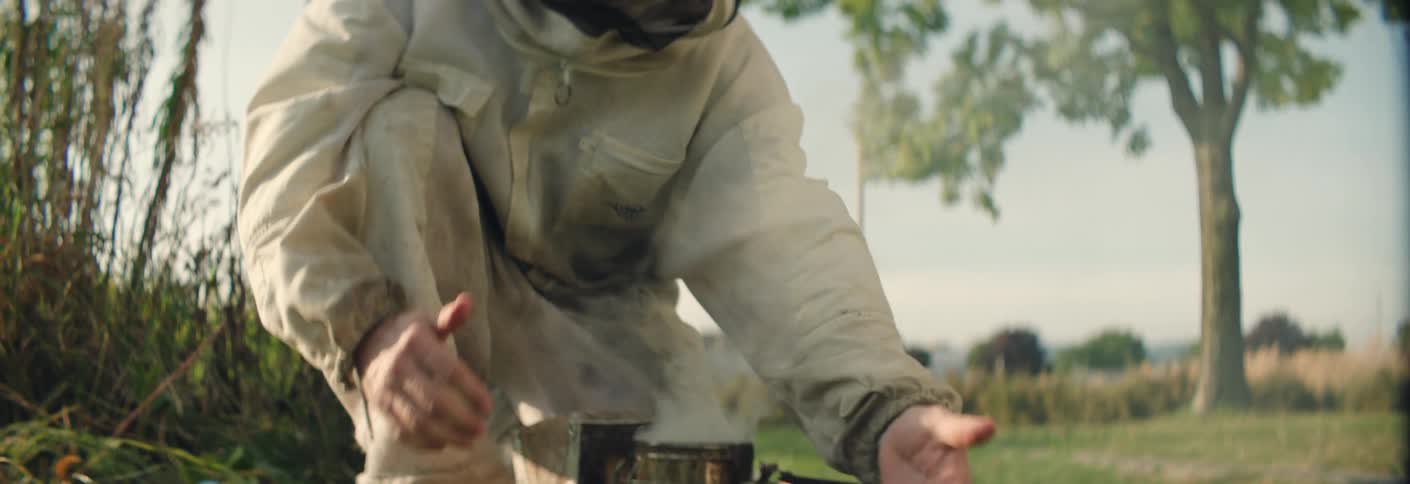 Beekeeper lights a metal smoker in a grassy field