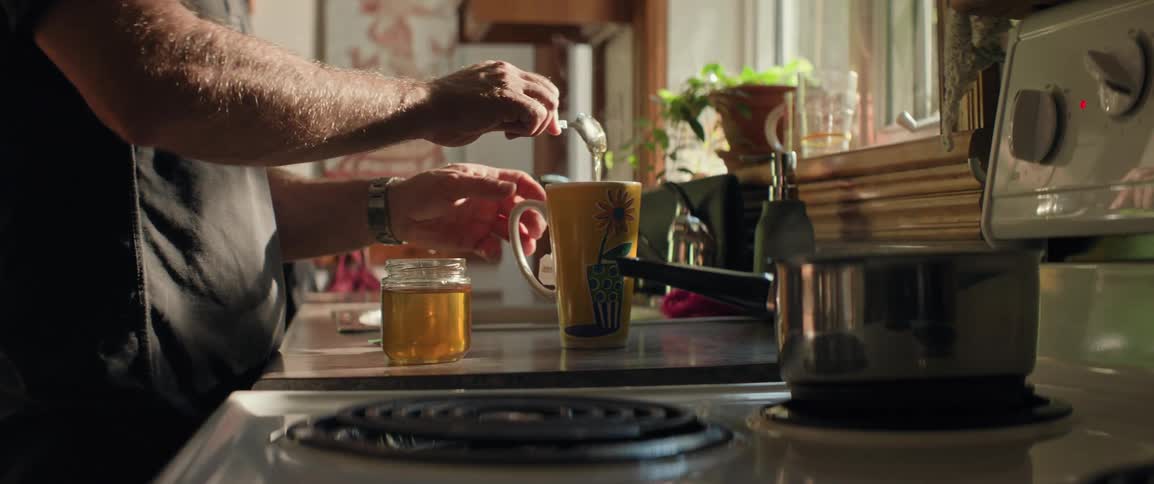 A person stirs honey into a mug of tea in a warmly lit kitchen with sunlight streaming through the window