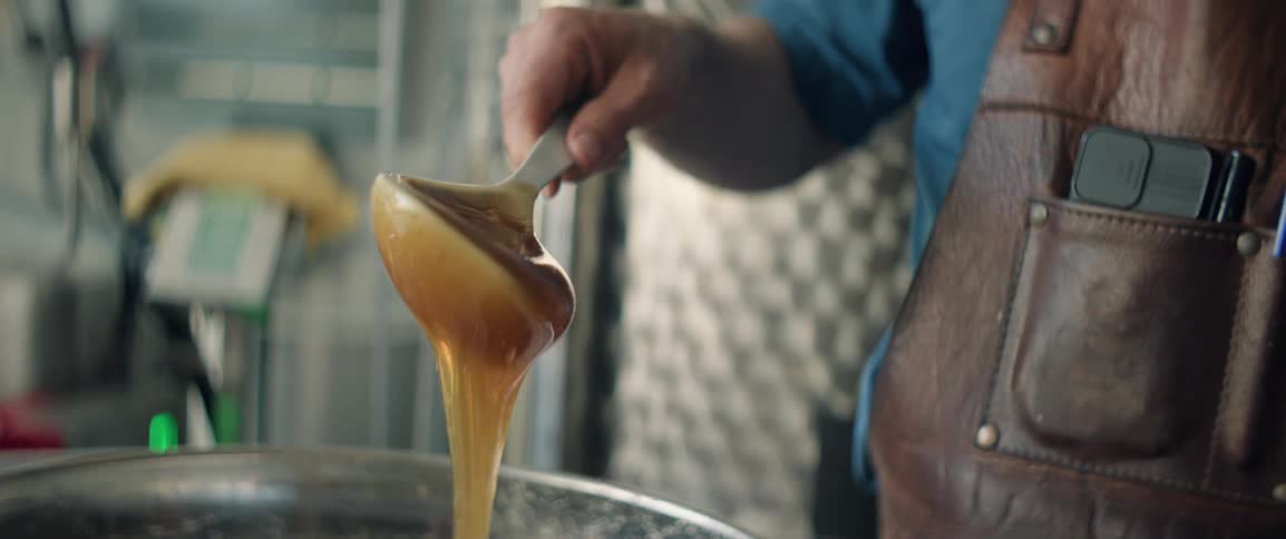 A man in an apron uses a large spoon to stir and then scoop honey